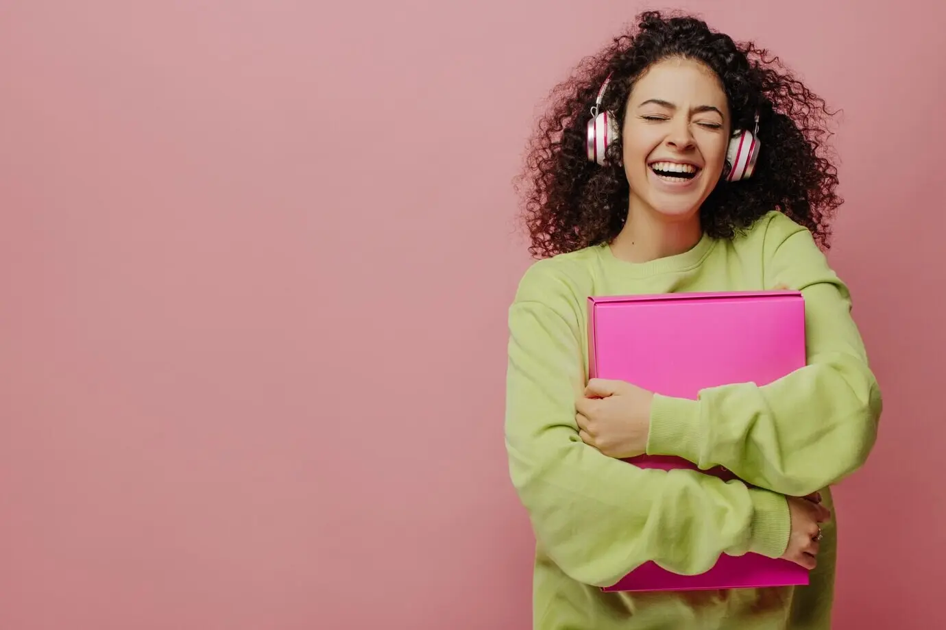 Jeune fille à la peau claire, brune aux cheveux bouclés, riant les yeux fermés en serrant une boîte contre sa poitrine; elle porte un casque audio et écoute sa musique préférée. Arrière-plan avec espace pour texte. Concept de profiter de l’instant.